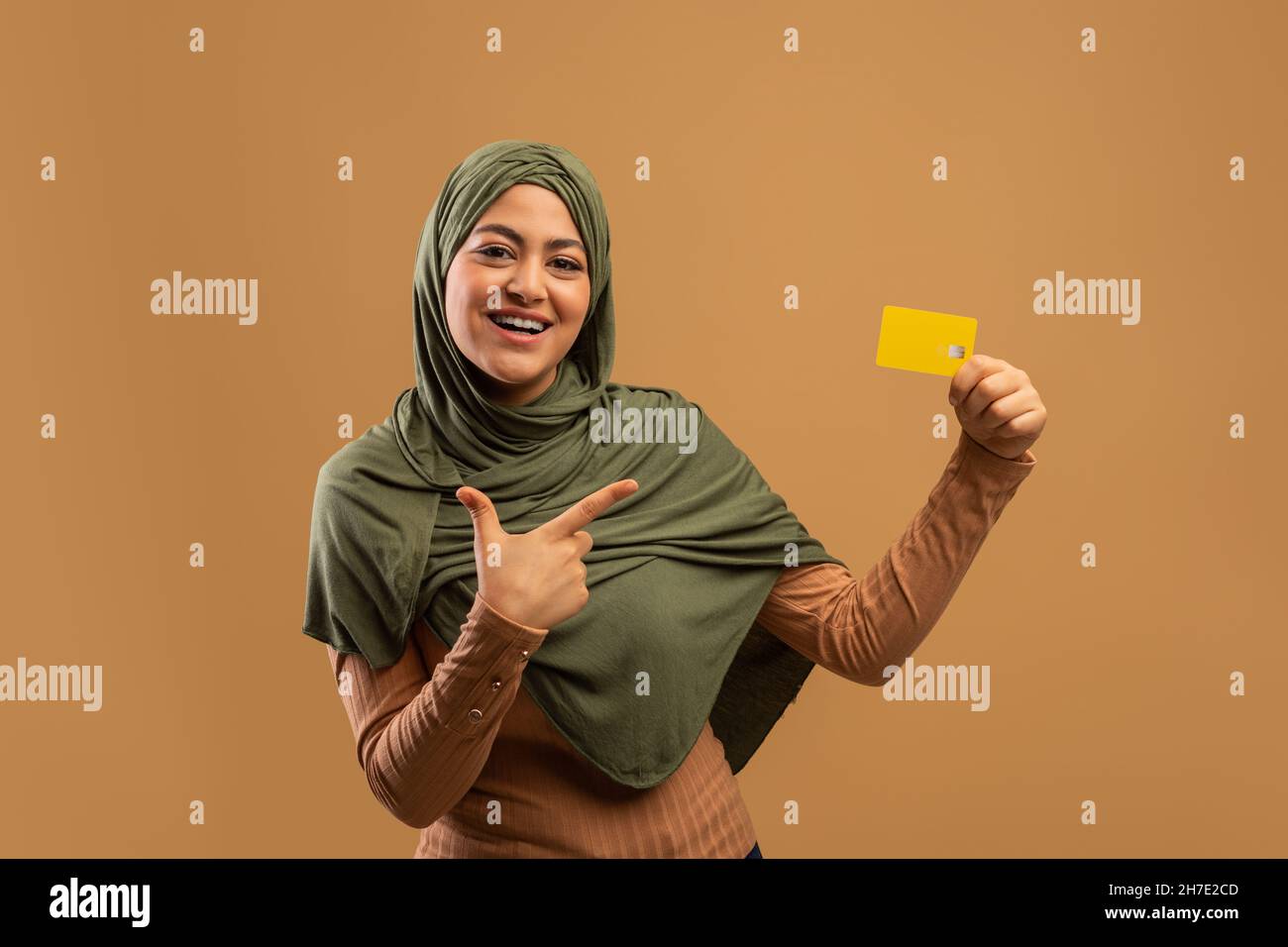 Kontaktloses Shopping-Konzept. Glückliche muslimische Dame zeigt auf Kreditkarte, empfiehlt Service, brauner Hintergrund Stockfoto