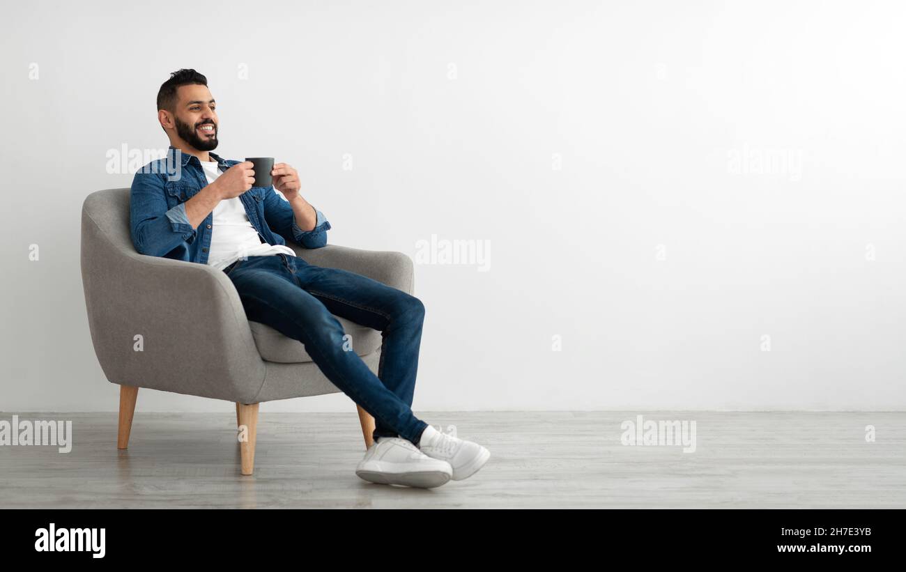 Ganzer Länge des jungen arabischen Mannes, der heißen Kaffee im Sessel gegen weiße Studiowand trinkt, Banner mit freiem Platz Stockfoto