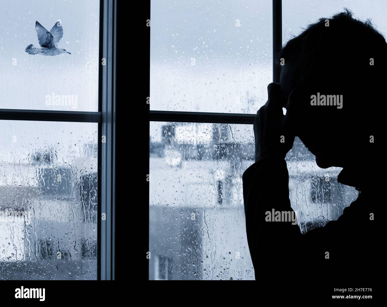 Mann mit der Hand am Kopf in der Nähe des Fensters an einem regnerischen Tag, an dem der Vogel vorbeifliegt. Konzept für Depressionen, Depressionen bei Männern, psychische Gesundheit, psychische Erkrankungen... Stockfoto