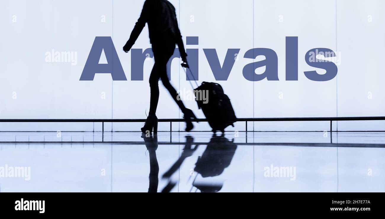Frau mit Gepäck auf Rädern/Koffer, die am Ankunftsschild am Flughafenterminal vorbeikommt. Stockfoto