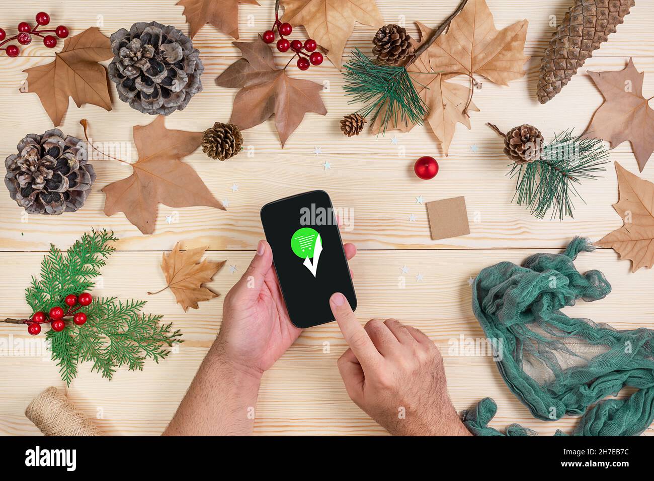 Draufsicht auf einen Holzhintergrund mit getrockneten Blättern und Weihnachtsmotiven mit den Händen eines Mannes, der nach Abschluss seines Online-Shoppins ein Handy in der Hand hält Stockfoto