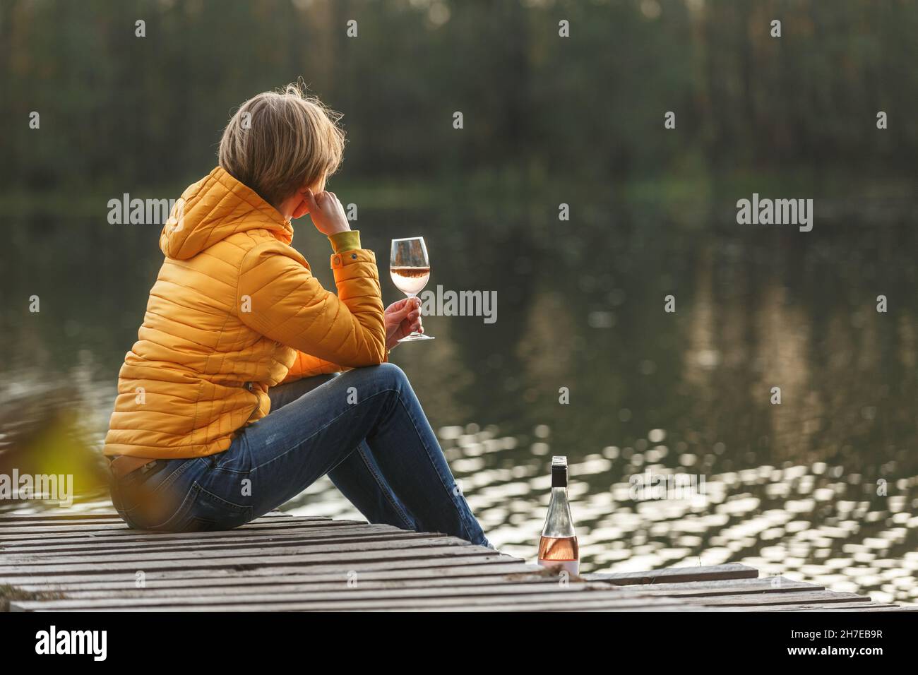 Frau in einer gelben Jacke, die sich auf einem hölzernen Pier auf einem See mit einem Glas Rosenwein entspannt und den Sonnenuntergang im Herbst alleine beobachtet. Genießen Sie die Natur, Entspannung und med Stockfoto