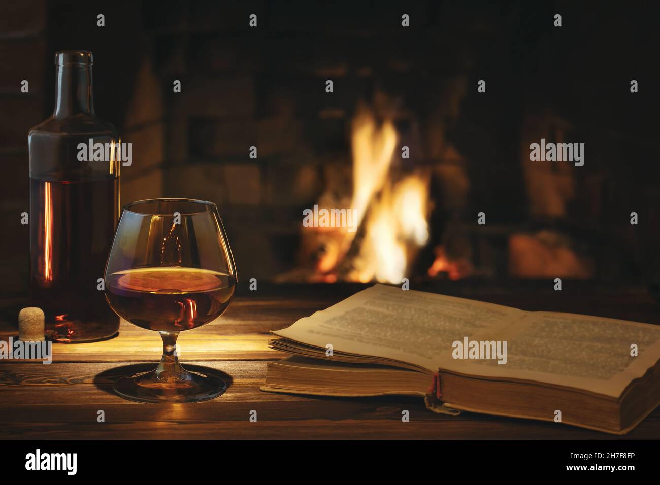 Ein Glas Cognac, eine Flasche und ein offenes altes Buch auf dem Tisch in der Nähe des brennenden Kamins. Ruhe- und Entspannungskonzept Stockfoto