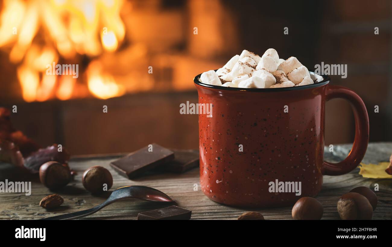 Kakao mit Salmellchen und Schokolade in einem roten Becher auf einem Holztisch in der Nähe eines brennenden Kamins. Stockfoto