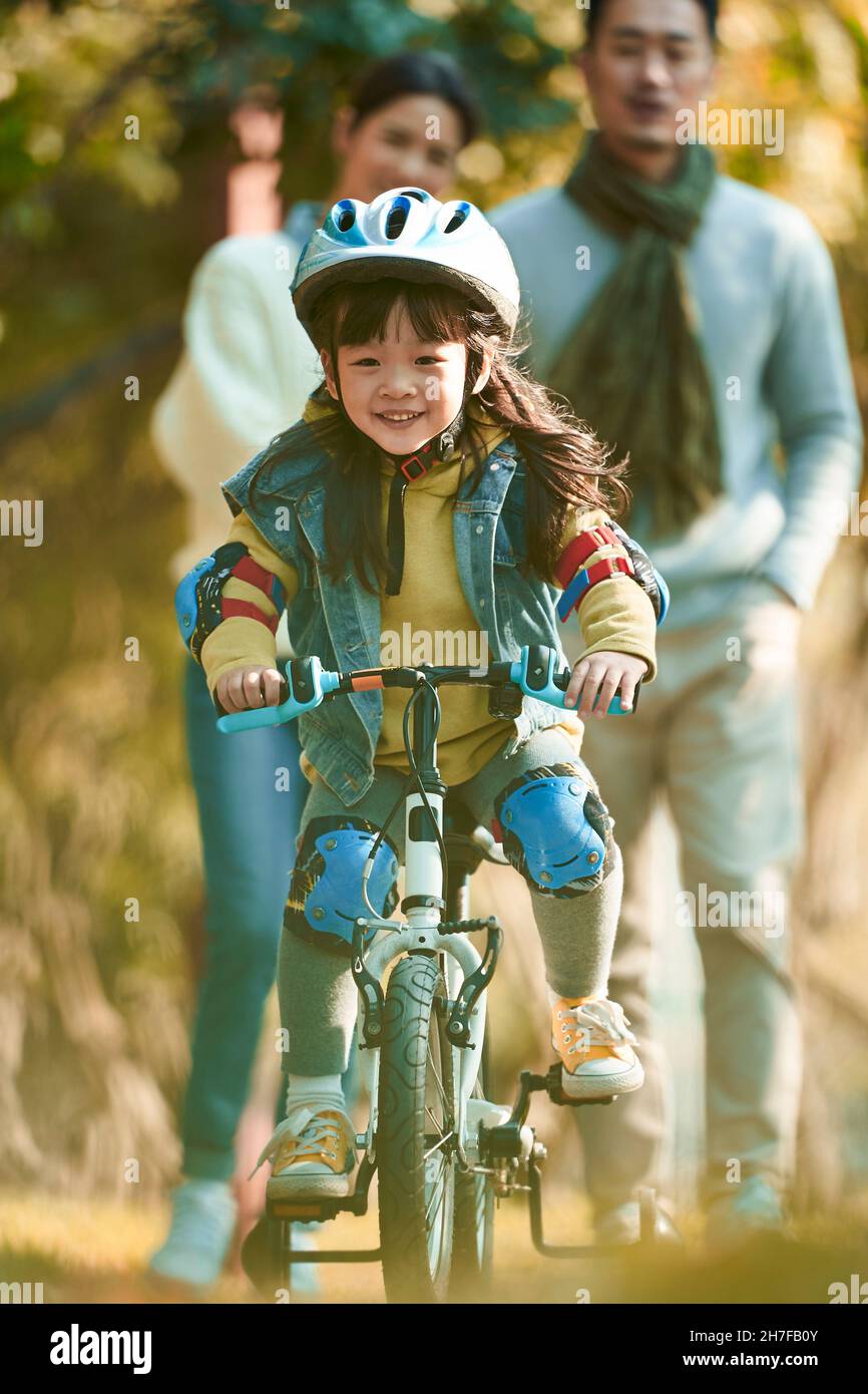 Kleine asiatische Mädchen mit Helm und voller Schutz Zahnräder Reiten Fahrrad im Stadtpark mit Eltern beobachten von hinten Stockfoto