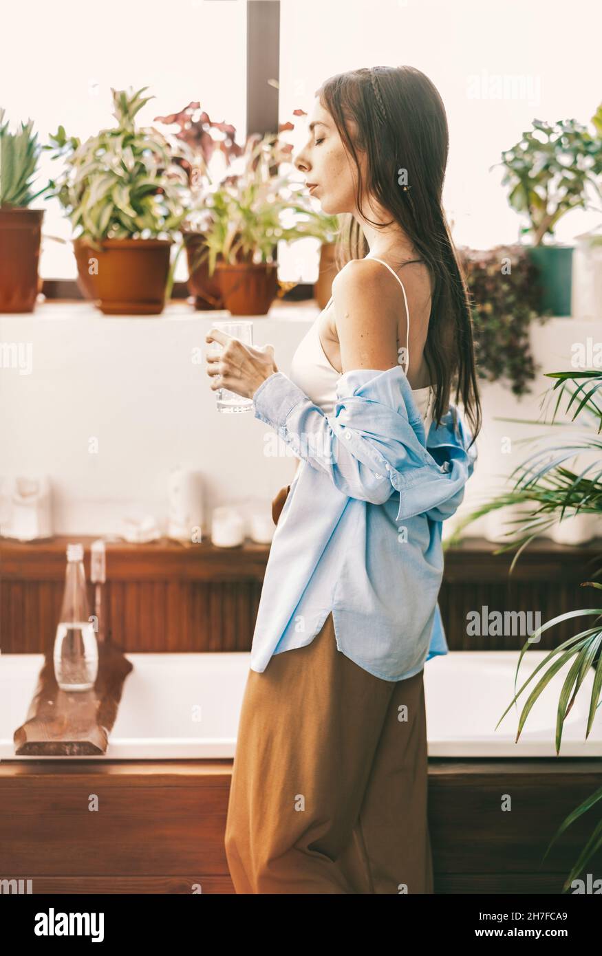 Ein Mädchen mit langen Haaren, in Hauskleidung, trinkt sauberes Wasser aus einem Glas im Badezimmer. Freies und ruhiges Mädchen beim Bad, Boho-Stil. Stockfoto