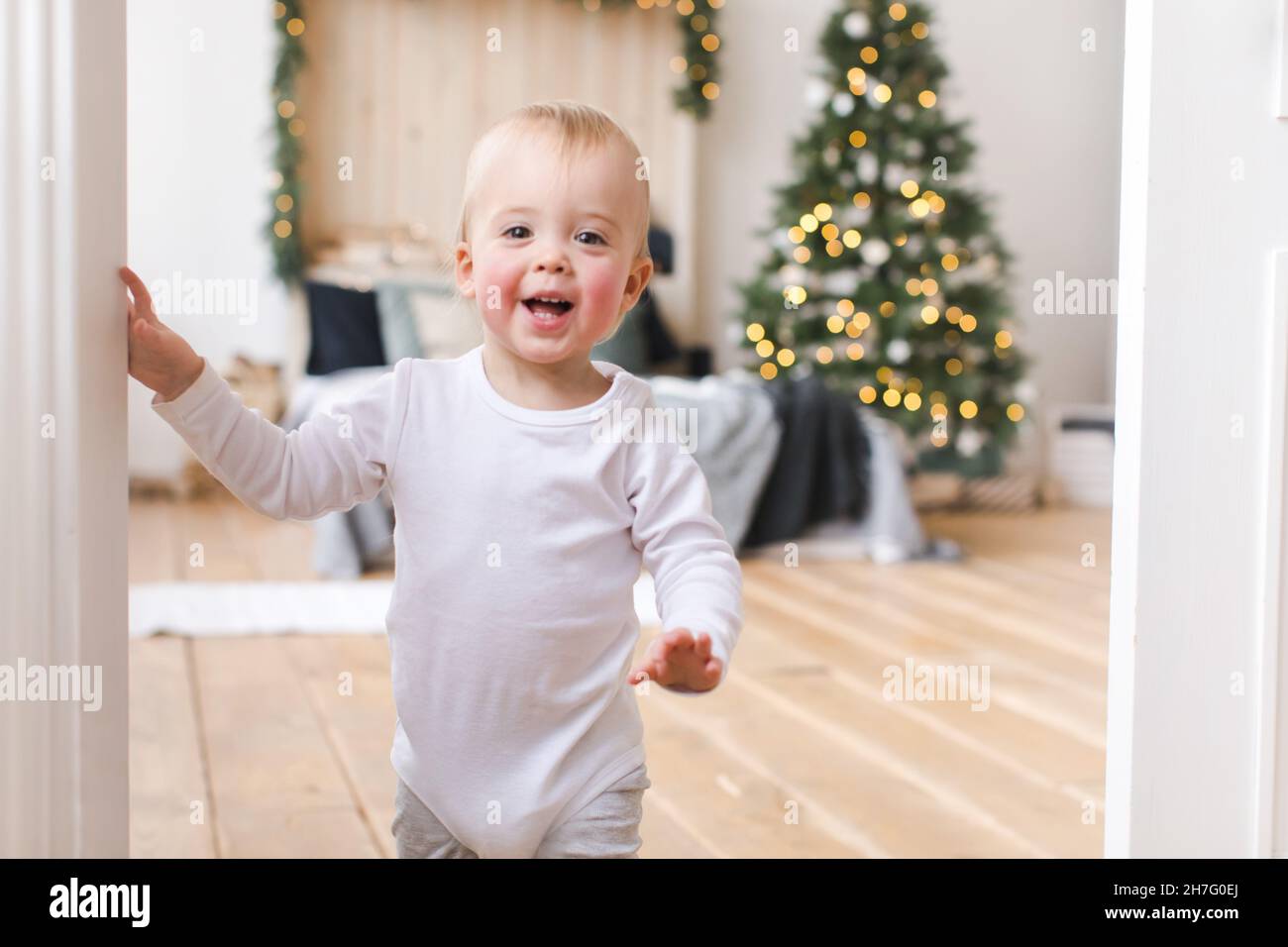 Entzückendes Kleinkind, das in den Türen steht und die Kamera auf dem Hintergrund des Weihnachtsbaums im Schlafzimmer anschaut. Stockfoto