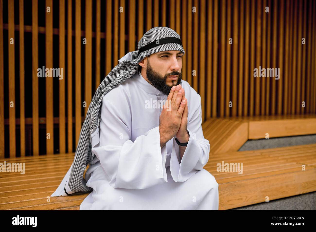 Junger Mann aus dem Nahen Osten in traditioneller Kleidung, der auf einer Holzbank sitzt Stockfoto