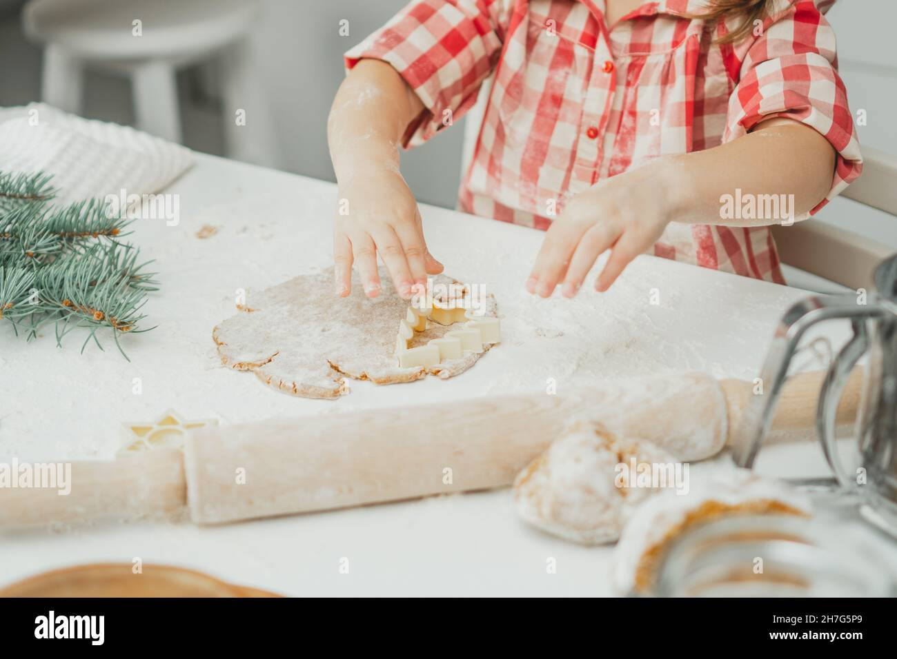 Teilweise verschwommene Hände eines kleinen Mädchens schneiden in der weißen Küche Weihnachtsbaum-Lebkuchenkekse aus gerolltem Teig aus. Das Kind bereitet Kekse auf Weiß zu Stockfoto
