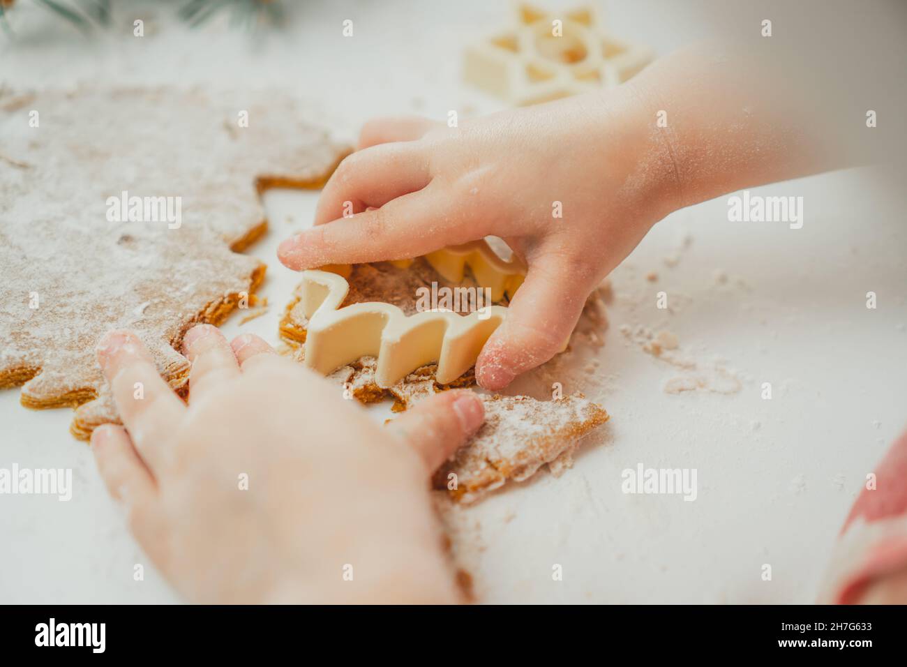 Teilweise verschwommene Hände eines kleinen Mädchens schneiden in der weißen Küche Weihnachtsbaum-Lebkuchenkekse aus gerolltem Teig aus. Das Kind bereitet Kekse auf Weiß zu Stockfoto