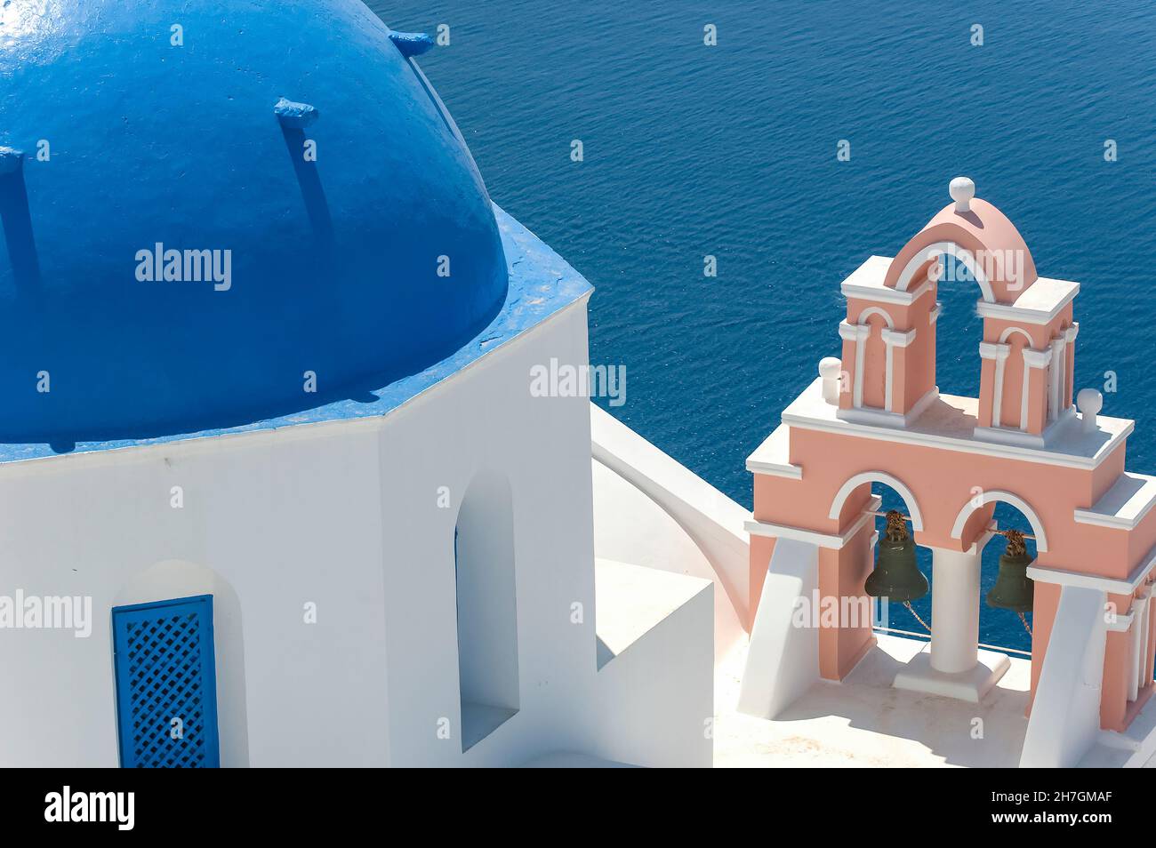 Nahaufnahme einer typischen weißen Kirche mit gebläutem Kuppeldach auf Santorini, Griechenland, mit farbenprächtigem rosafarbenem Glockenturm vor dem Hintergrund des blauen Wassers von Th Stockfoto