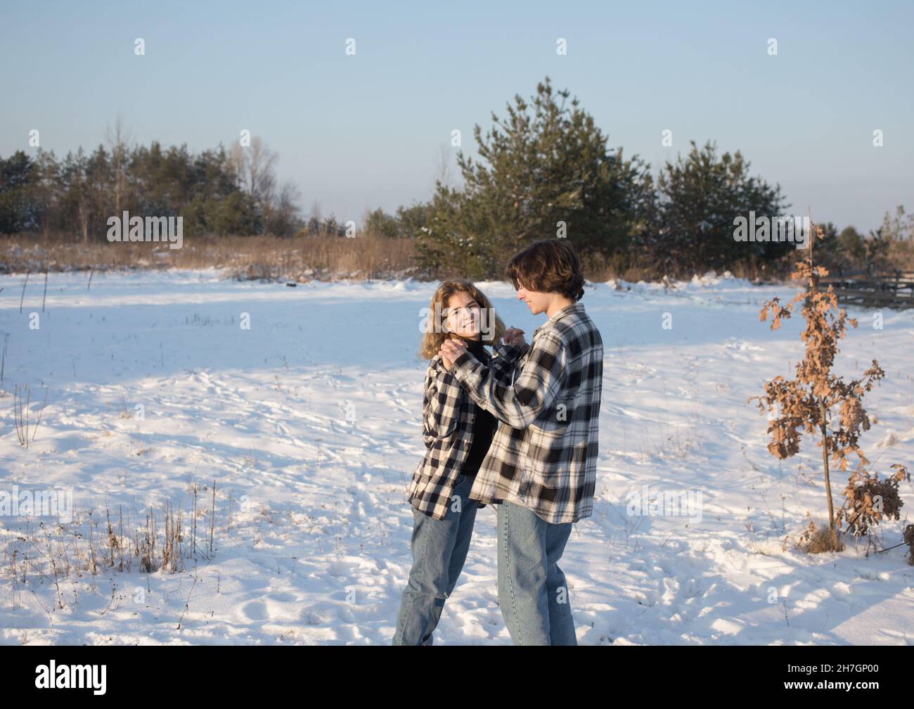 Ein fröhliches, verliebtes Teenager-Mädchen und ein etwa 16-17-jähriger Kerl in karierten Hemden und Jeans stehen im Schnee, umarmen sich und lachen fröhlich. Die Conce Stockfoto