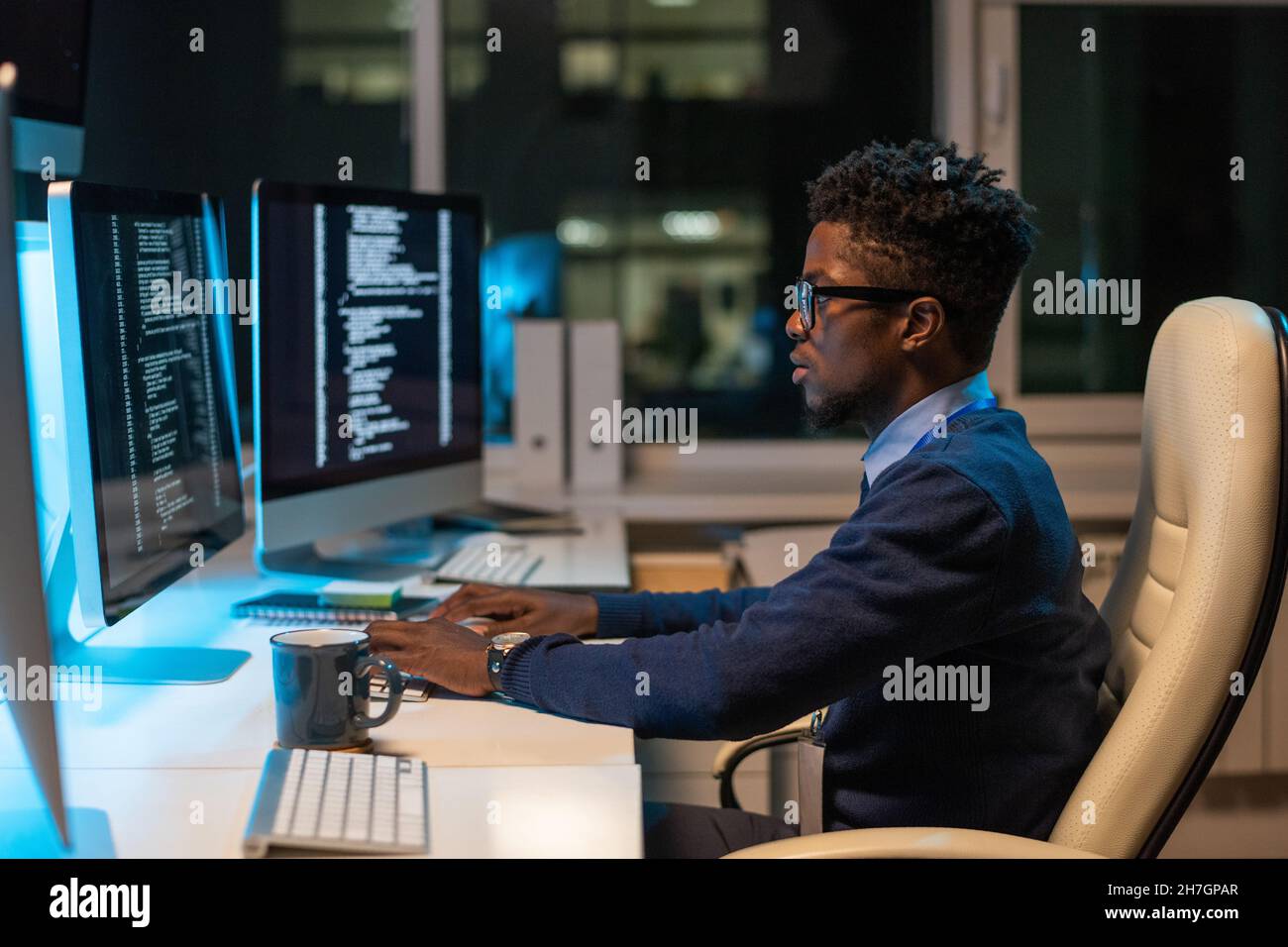 Junger IT-Ingenieur afrikanischer Herkunft, der mit kodierten Daten arbeitet, während er im Büro im Sessel vor dem Computer sitzt Stockfoto