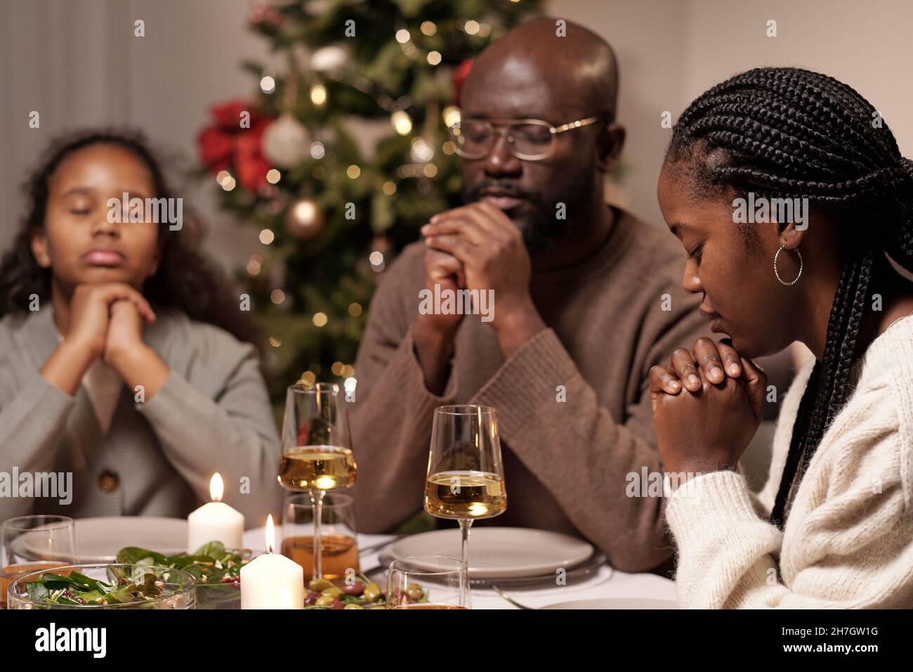 Die junge Afrikanerin, ihr Mann und ihre süße Tochter beten vor dem Abendessen mit den Händen am Kinn zusammen Stockfoto