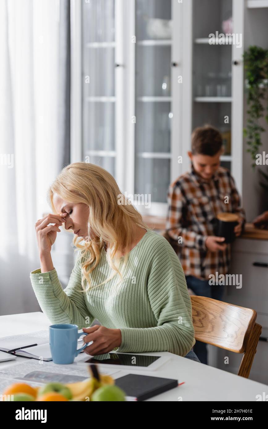 Müde Frau mit geschlossenen Augen sitzt mit einer Tasse Tee und digitale Tablette mit leerem Bildschirm in der Nähe verschwommen Sohn in der Küche Stockfoto