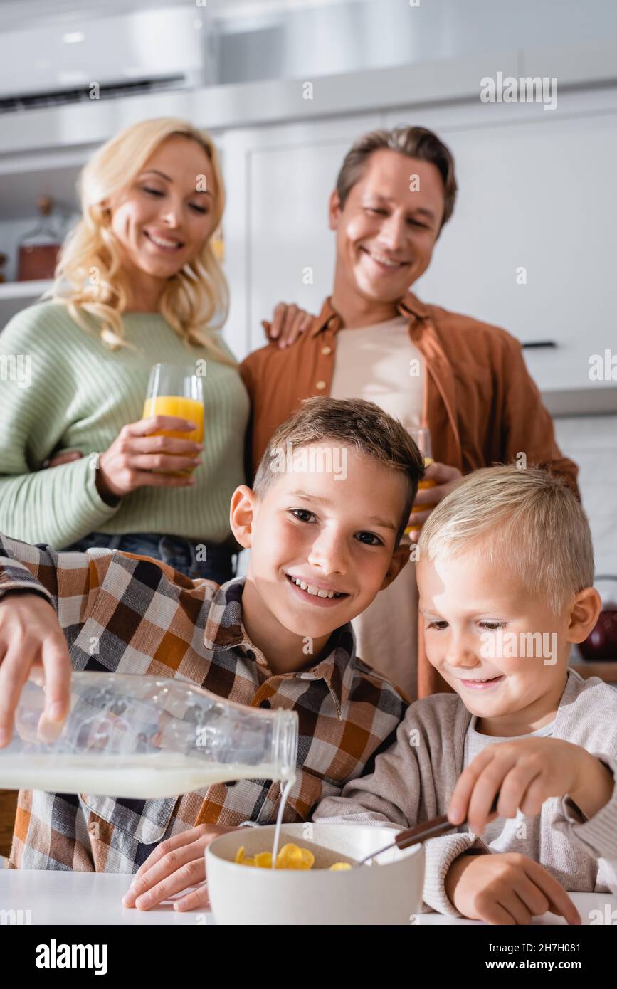 Lächelndes Kind, das die Kamera anschaut, während es Milch mit leckeren Cornflakes in die Schüssel gießt Stockfoto