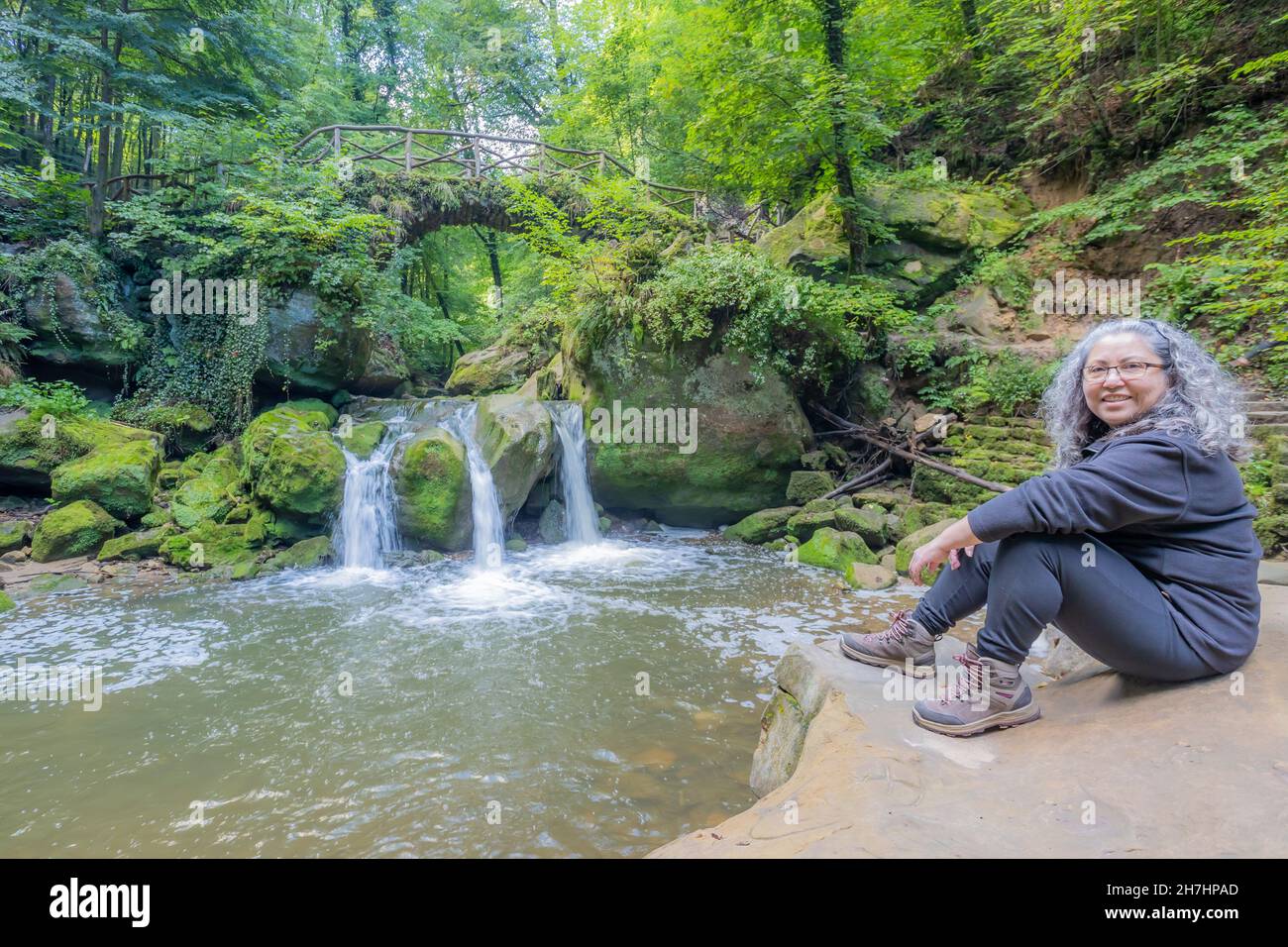 Wandertouristin auf Felsen sitzend, Schiessentümpel Wasserfall am Black Ernz Fluss und üppige grüne Wildvegetation im Hintergrund, gräuliches Haar, lo Stockfoto