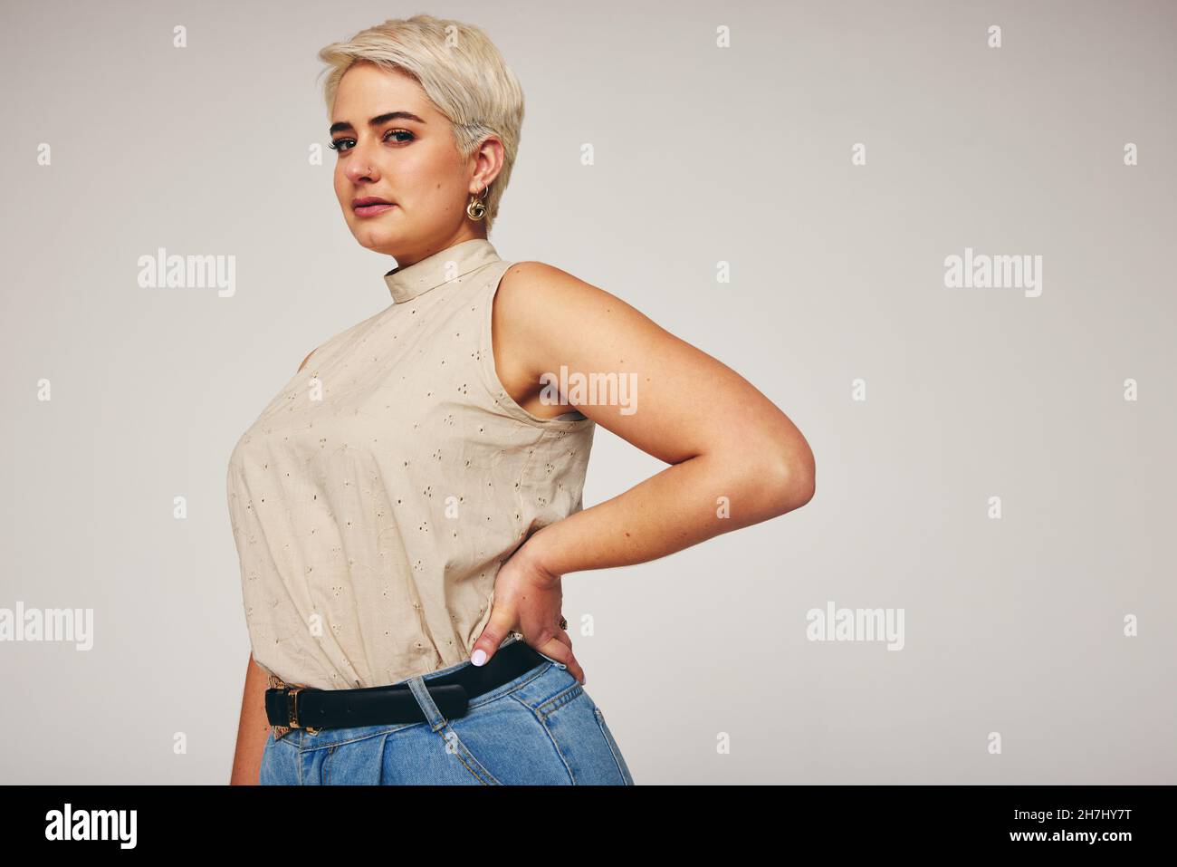 Blonde Frau, die selbstbewusst auf die Kamera schaut, während sie vor grauem Hintergrund steht. Modische Frau mit mittleren Erwachsenen, die Denim-Jeans in einer Stu trägt Stockfoto