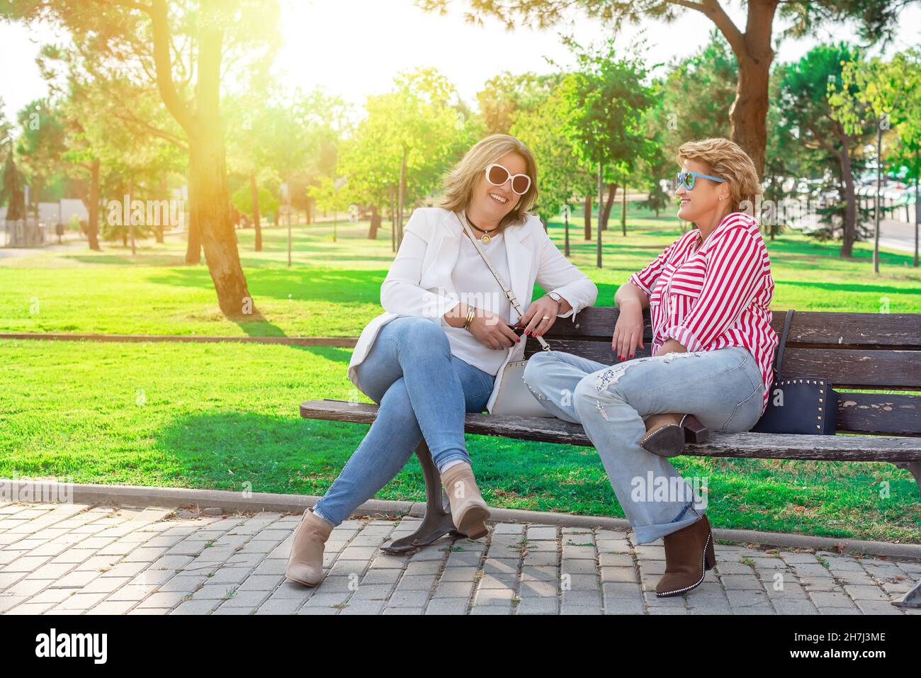 Zwei kaukasische Frauen mittleren Alters sitzen auf einer Parkbank und lachen und plaudern lebhaft. Stockfoto