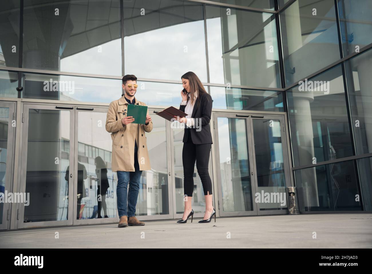 Die Chefin und ihr männlicher Mitarbeiter lächeln zusammen, während sie vor dem Unternehmen am Telefon telefonieren Stockfoto