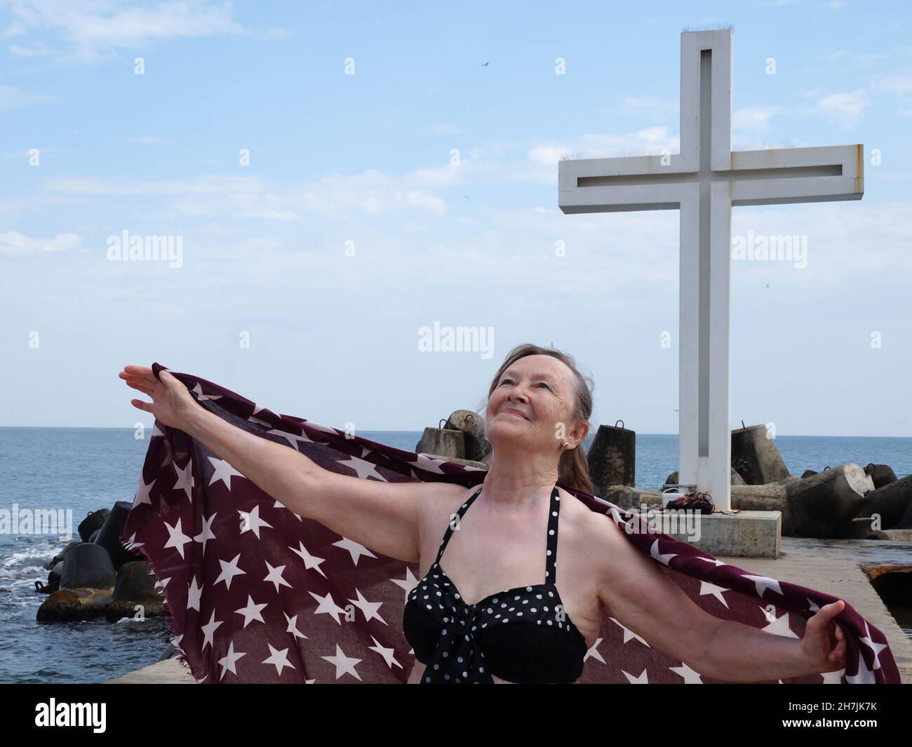 Eine ältere lächelnde Frau, die einen Schal im Wind mit offenen Armen an der Küste vor dem Hintergrund eines großen Kreuzes hält. Stockfoto
