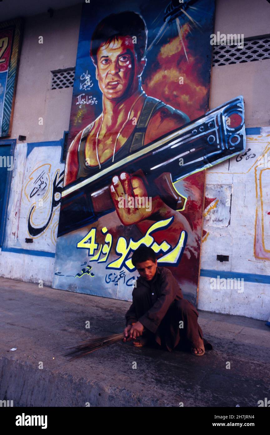 Ein junger afghanischer Junge fegt den Boden sauber, vor einem überlebensgroßen Poster des beliebten Action-Films Rambo, im Kissa Khuwani Bazaar in Pesha Stockfoto