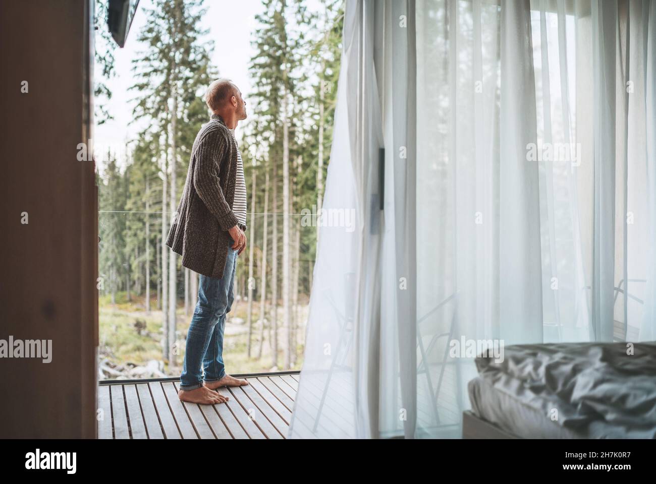 Ein Mann gekleidet offene Strickjacke auf einem Waldhaus Balkon stehen und genießen die frische Luft mit Natur Wald Pinien Blick. Außerhalb Von Scandina Stockfoto