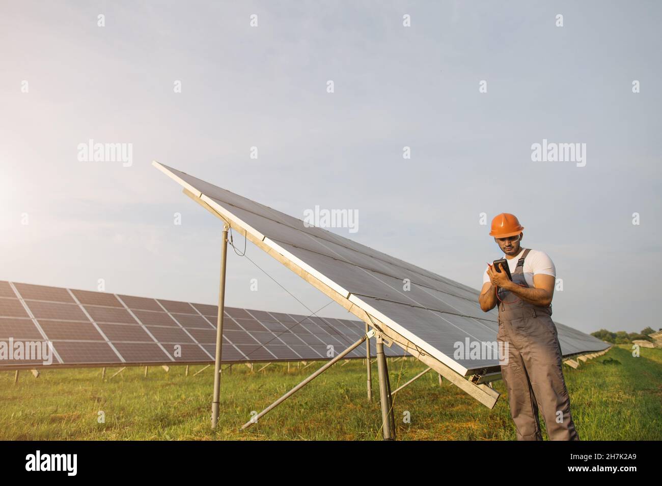 Indischer Arbeiter in Uniform, Schutzhelm und Brille mit Multimeter, während er die Arbeit von Solarmodulen untersucht. Konzept von Menschen, Wartung und grüner Energie. Stockfoto