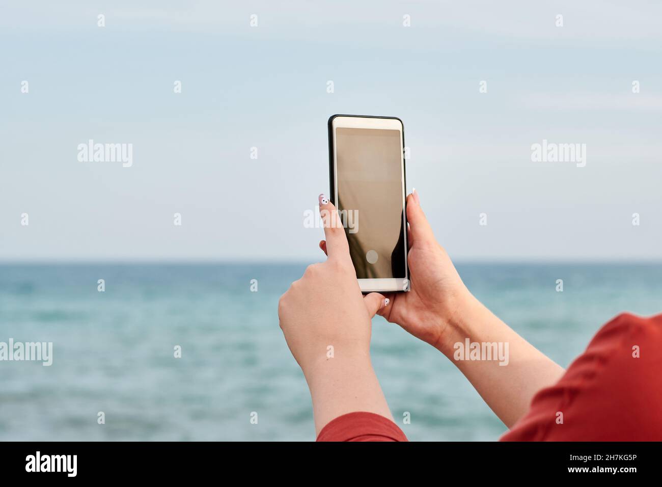 Hände einer Person, die ein Handy mit dem Meer im Hintergrund hält Stockfoto