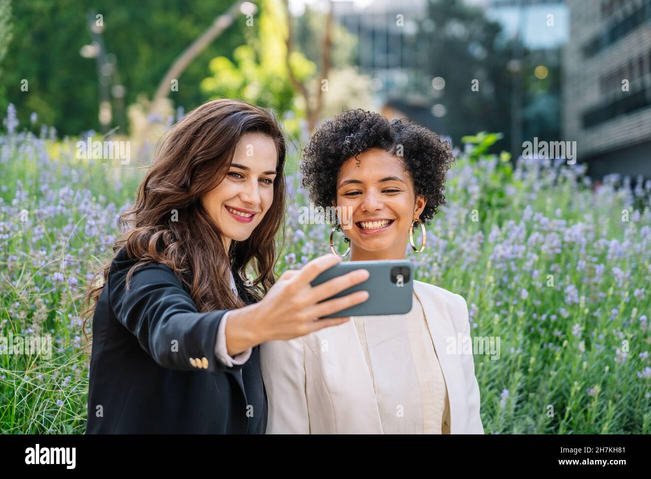 Fröhliche junge multirassische Freundinnen in eleganten Outfits lächeln, während sie am sonnigen Tag im blühenden Park auf dem Smartphone Selbstporträt machen Stockfoto