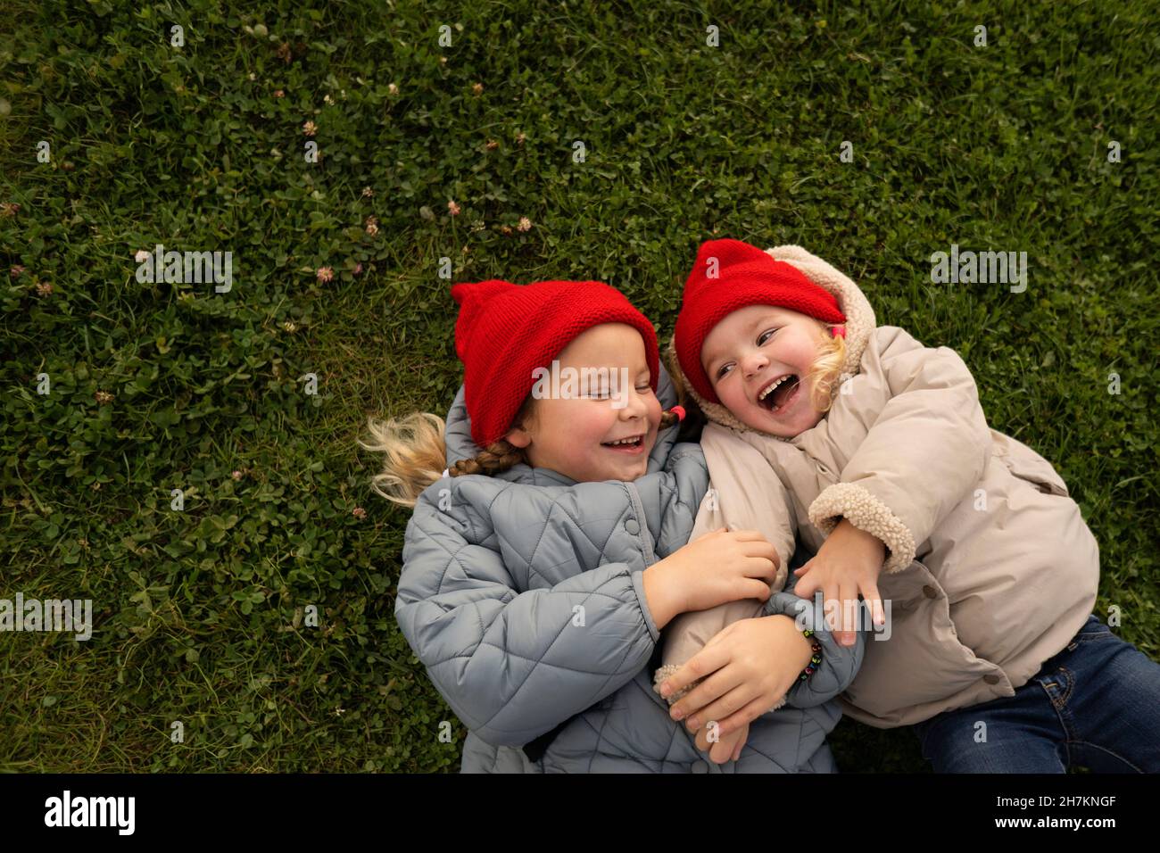 Mädchen in Strickmütze kitzelt Schwester, während sie auf dem Rasen auf dem Spielplatz liegt Stockfoto