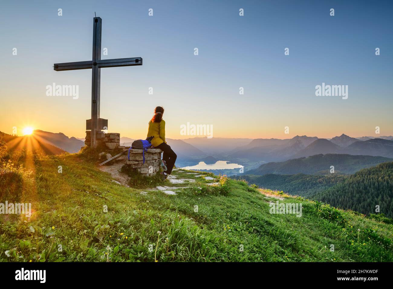 Wanderweibchen, die bei Sonnenaufgang am Gipfelkreuz auf dem Berggipfel sitzt, Kärnten, Österreich Stockfoto