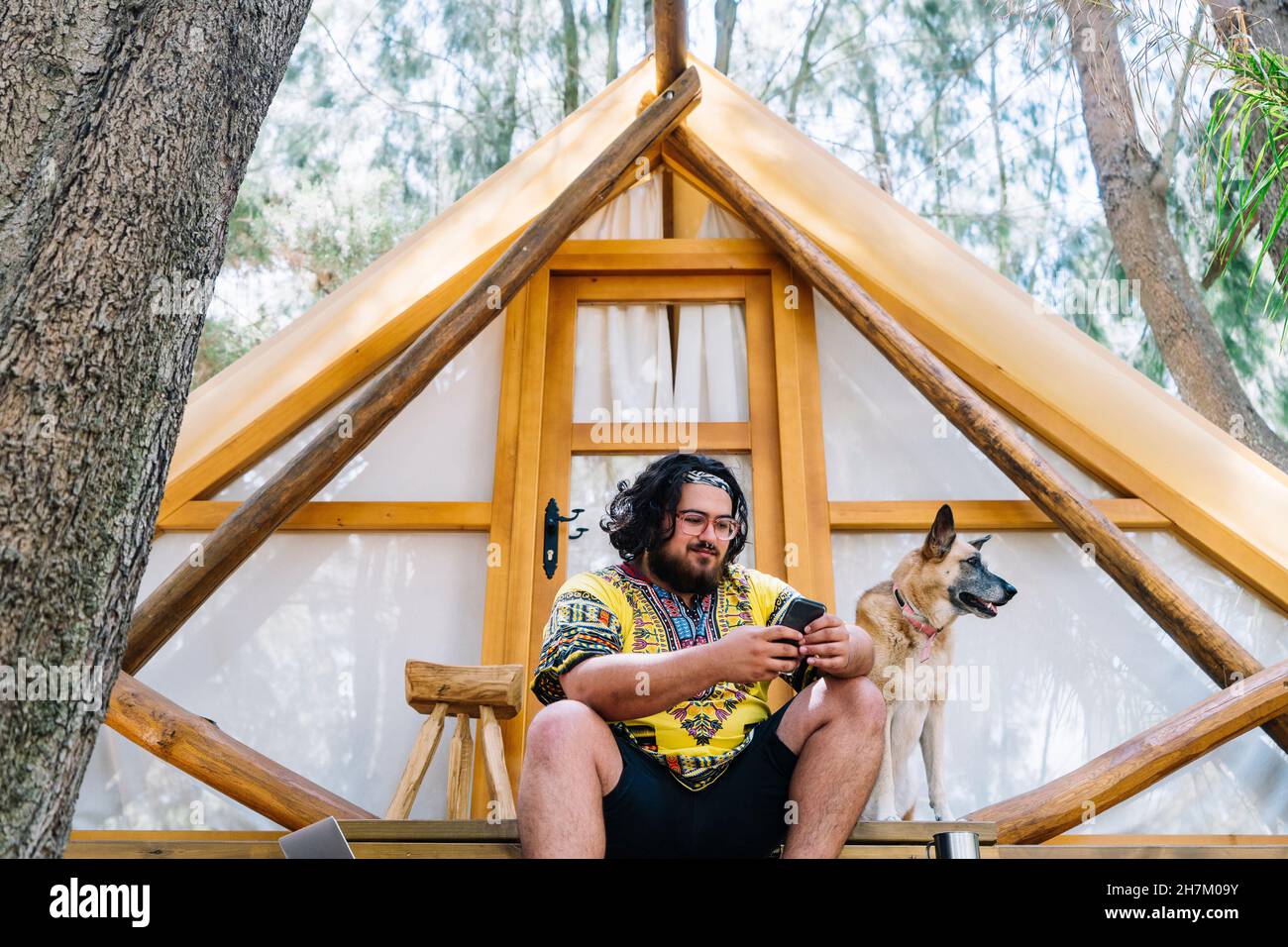 Mann, der am Eingang des Zeltes ein Mobiltelefon mit Hund benutzt Stockfoto