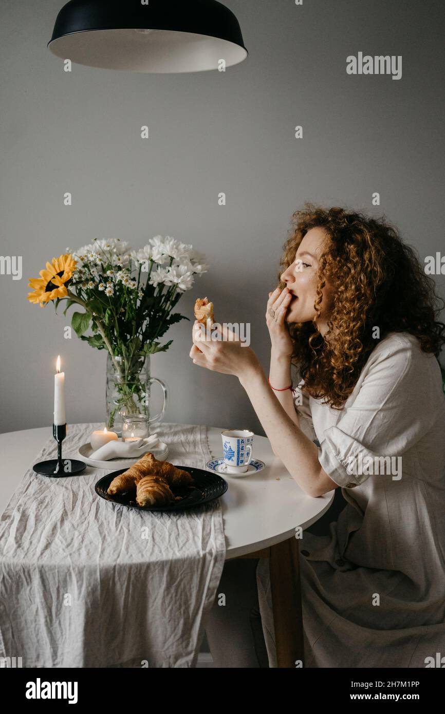 Frau, die Croissant am Tisch im Wohnzimmer isst Stockfoto