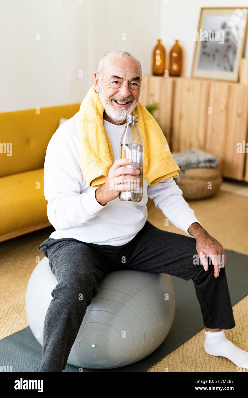 Mann mit Wasserflasche sitzt zu Hause auf dem Fitnessball Stockfoto