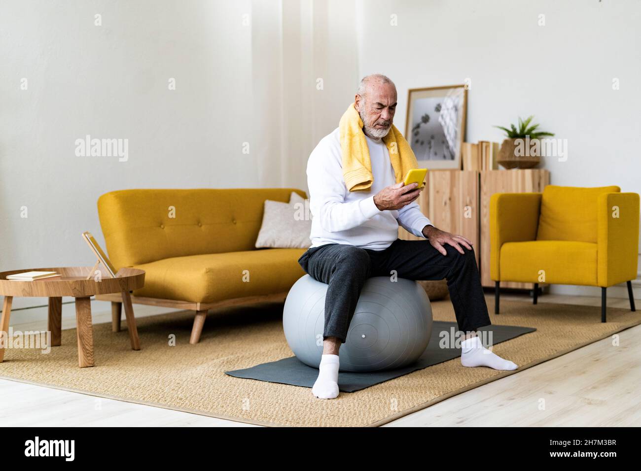 Ein älterer Mann mit Serviette um den Hals sitzt zu Hause auf dem Fitnessball Stockfoto