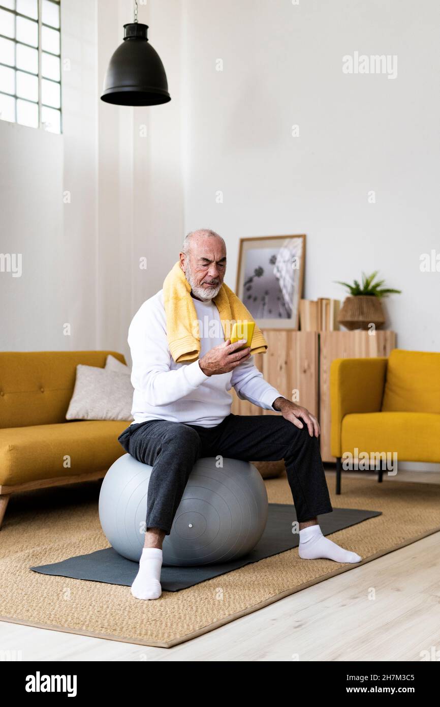 Älterer Mann, der im Wohnzimmer ein Mobiltelefon auf dem Fitnessball benutzt Stockfoto