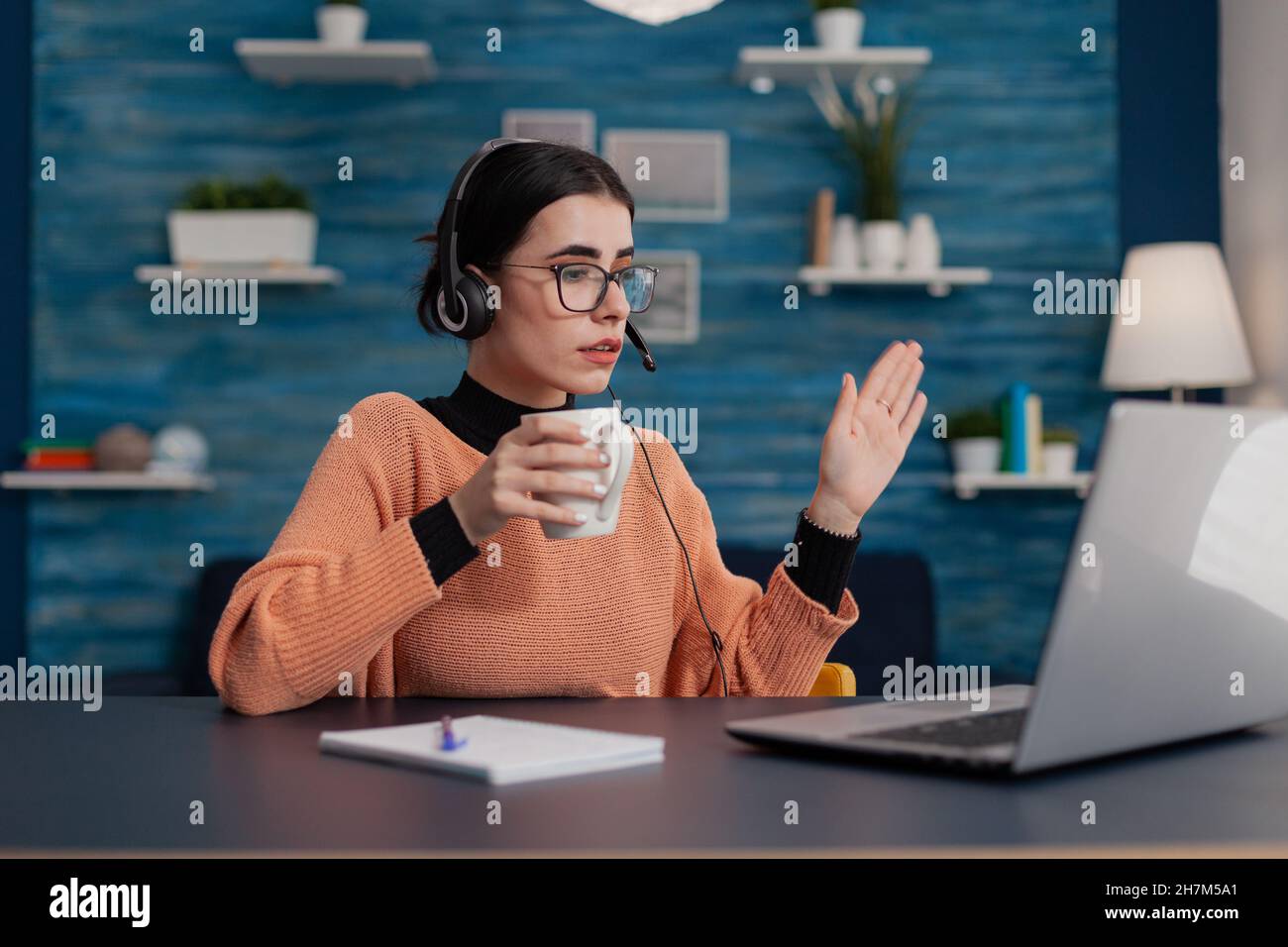 Schüler mit Brille winkt in Laptop-Videocall lässig hallo. Person mit Kopfhörern, die die Hand hebt, in einer Videoanruf-Vorlesung aus der Ferne. College Mädchen attendig online-Klasse aus dem Home Office. Stockfoto
