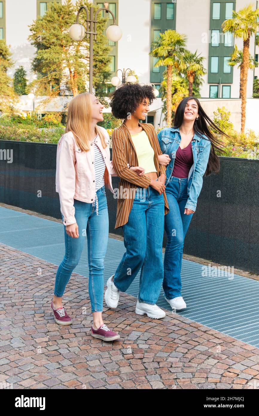 Ein ganzer Körper junger multirassischer Freundinnen in Jeans, die sich fröhlich unterhalten und lachen, während sie auf einem gepflasterten Gehweg in der Sommerstadt zusammen spazieren Stockfoto