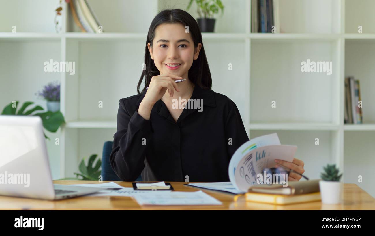 Attraktive asiatische junge Geschäftsfrau, die an ihrem Schreibtisch sitzt, mit den Augen auf der Kamera und der Hand auf dem Kinn. Stockfoto