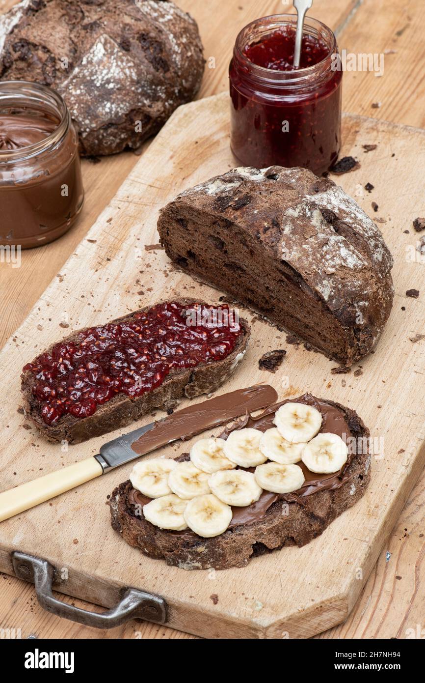 Chocolate Sourdough Bread mit Schokolade-Haselnuss-Aufstrich, Bananenscheiben und Himbeermarmelade auf einem Holzbrett Stockfoto
