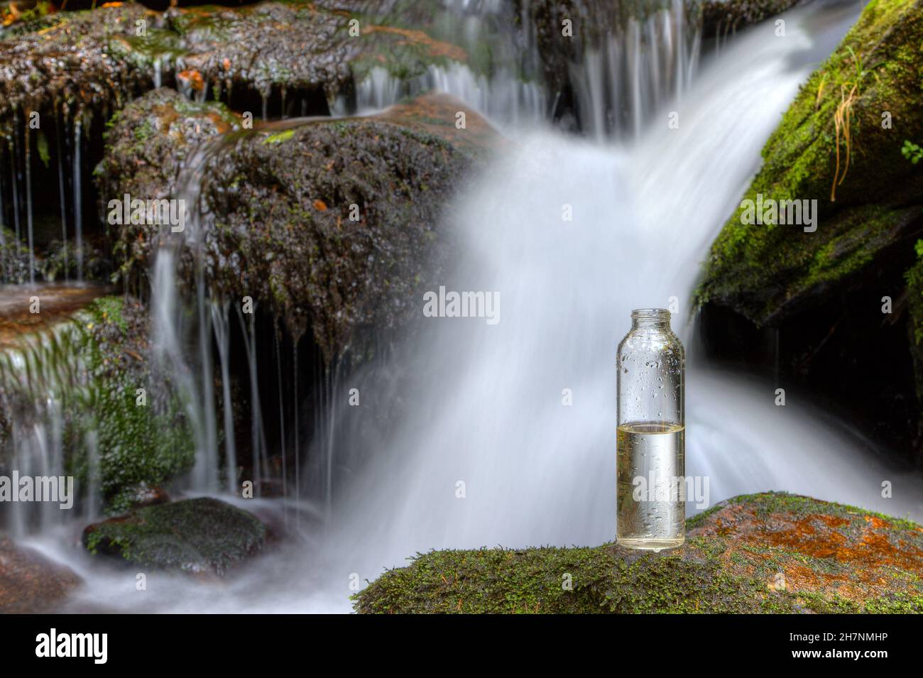 Eine halbvolle Flasche Trinkwasser steht auf einem Stein vor einem Wasserfall. Wird uns bald das Wasser ausgehen? Stockfoto