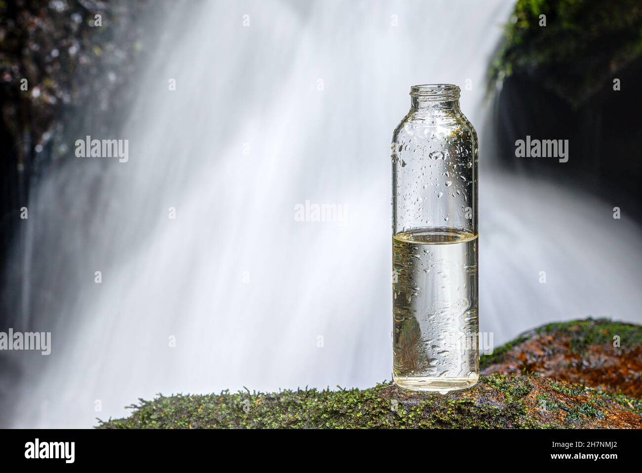 Eine halbvolle Flasche Trinkwasser steht auf einem Stein vor einem Wasserfall. Wird uns bald das Wasser ausgehen? Stockfoto