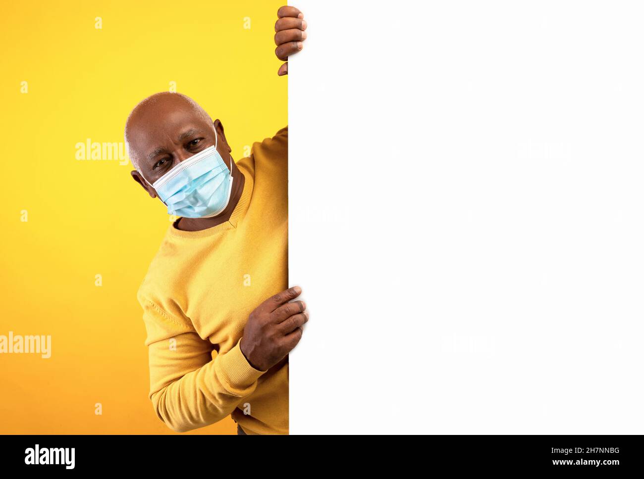 Älterer schwarzer Mann, der hinter einem leeren Banner herausschaut, eine Gesichtsmaske trägt, für Ihr Produkt oder Ihre Dienstleistung wirbt, ein Modell Stockfoto