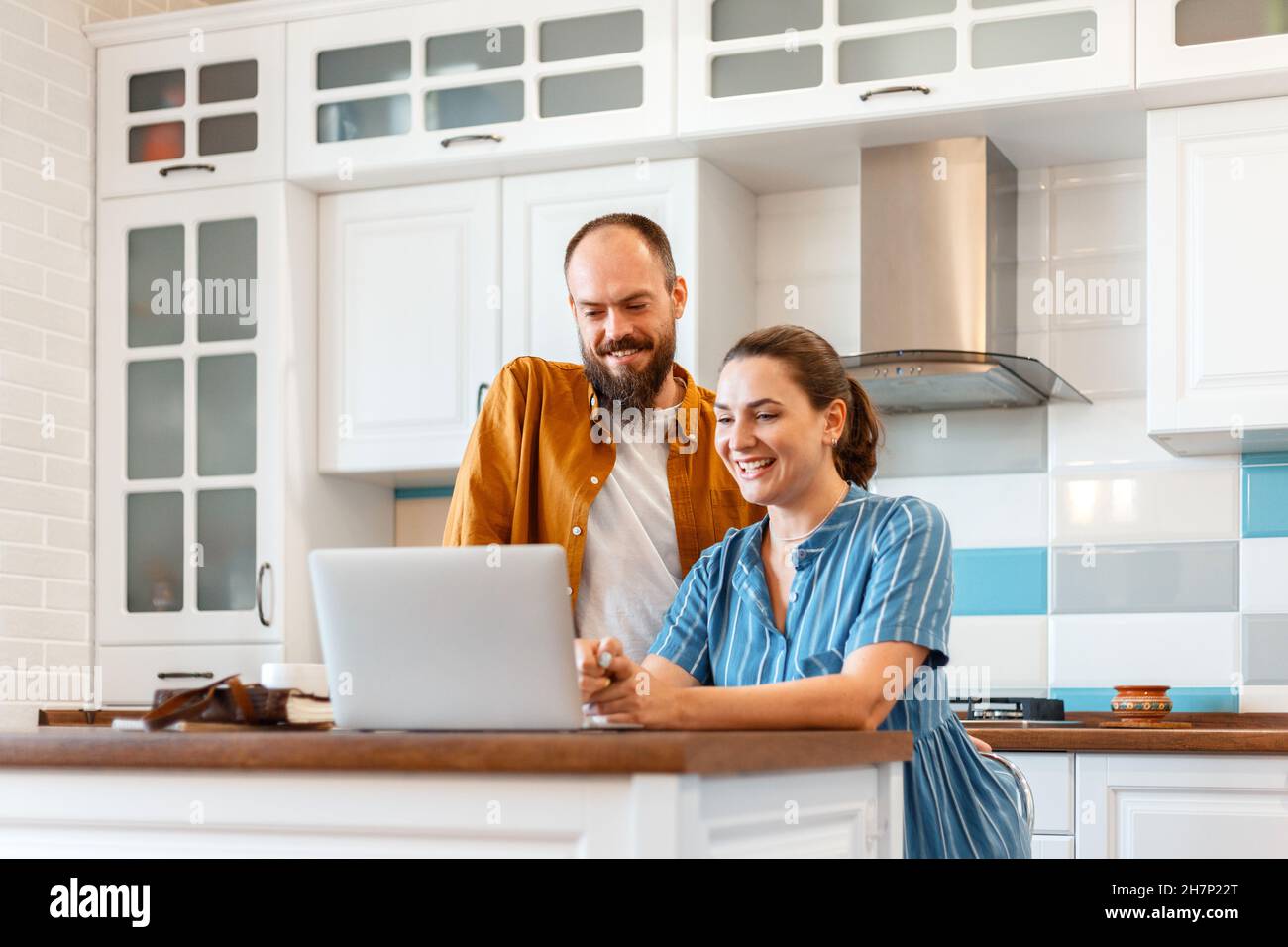 Lächelndes junges Ehepaar, das zu Hause in der Küche per Videoanruf mit einem Laptop spricht. Frau und Mann freuen sich über gute Nachrichten aus der E-Mail in zu hören Stockfoto