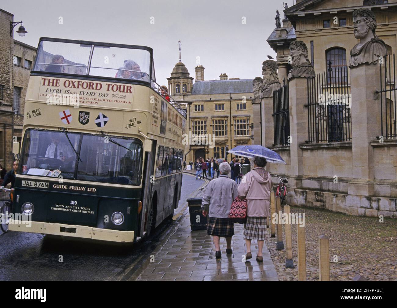 Zwei ältere Frauen mit Regenschirm und einem Touristenbus an einem regnerischen Tag in einer breiten Straße im Zentrum von Oxford, Großbritannien Stockfoto