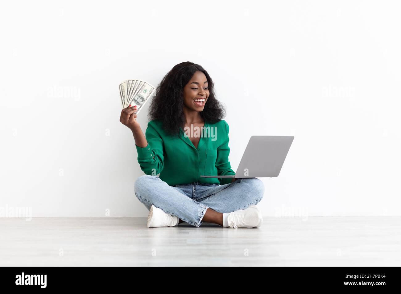 Glückliche afroamerikanische Dame spielt online, hält Laptop und Geld Stockfoto