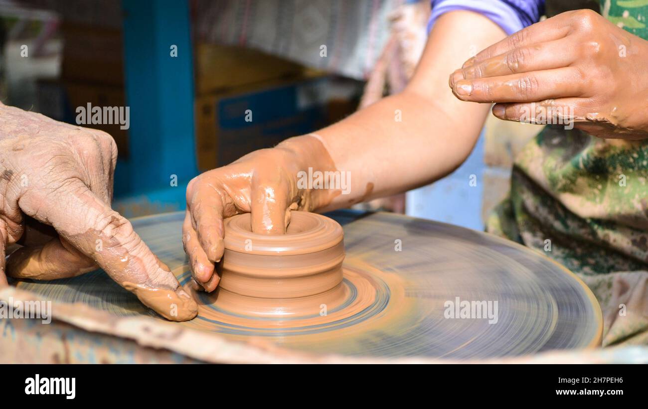 Die Hände eines alten Lehrers und eines jungen Studenten in Ton. Das Töpferrad dreht den Ton unter den Handflächen. Handgefertigt, mit natürlichen Materialien gearbeitet Stockfoto