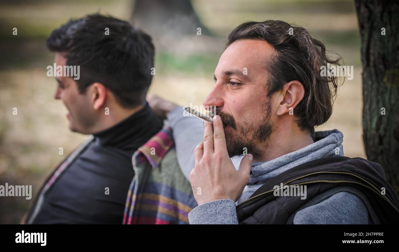Zwei entspannte und lächelnde, glückliche junge Freunde, die Cannabis-Marihuana-Ganja oder Haschisch-Joint im öffentlichen Park im Freien rauchen. Zwei Männer rauchen thc-grünes Gras t Stockfoto