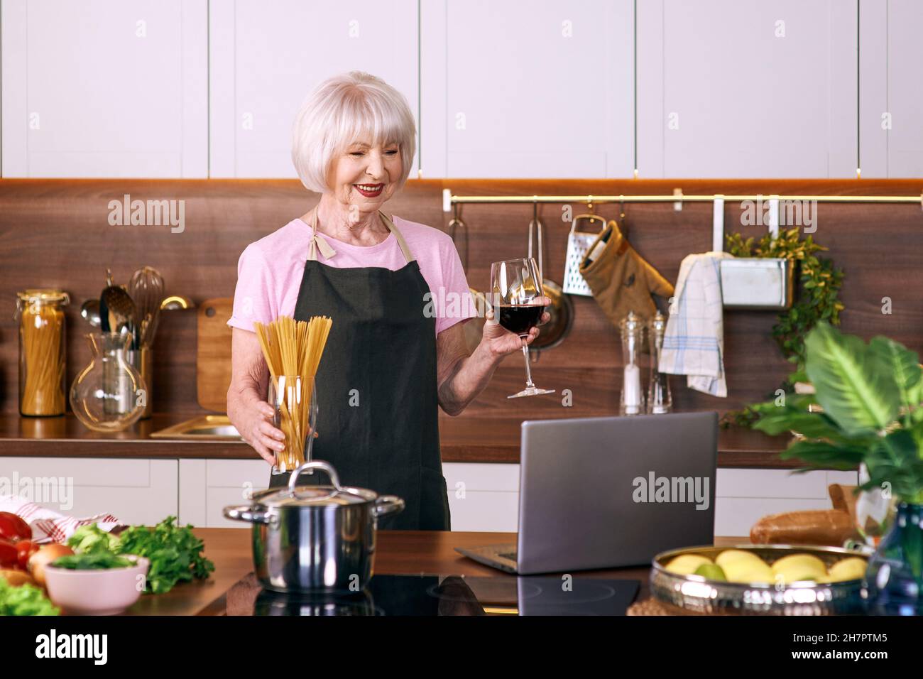 Ältere fröhliche Frau kocht in der modernen Küche mit einem Laptop. Essen, Bildung, Lifestyle-Konzept Stockfoto